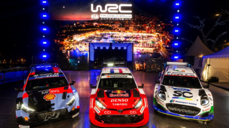 Three rally cars sit on a promotional stage in front of WRC logo and branding at the season opener in Monte Carlo. The cars are red, black and white.