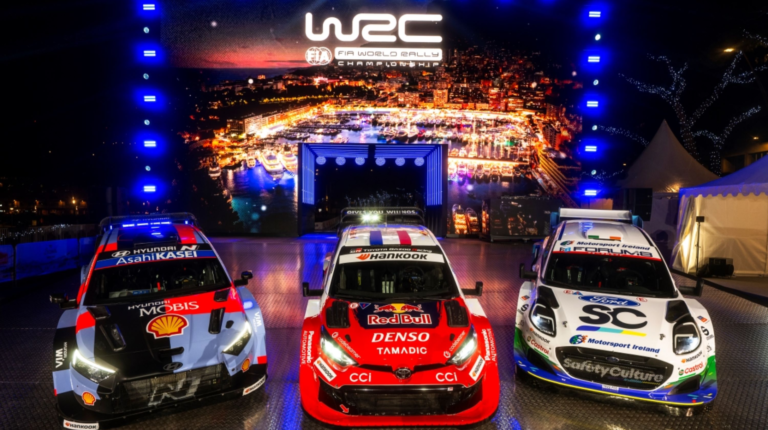 Tree rally cars sit on a promotional stage in front of WRC logo and branding at the season opener in Monte Carlo. The cars are red, black and white.