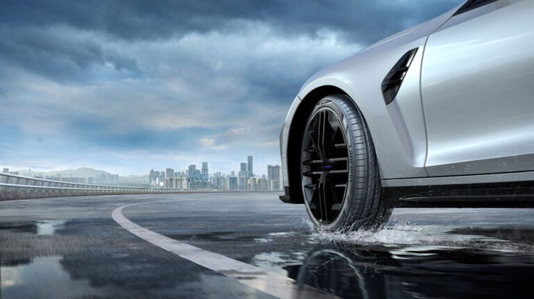 The Laufenn S Fit 2 summer tire on a grey vehicle driving on a water-covered road outside of a city.