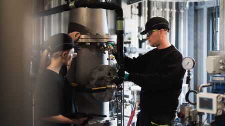 A manufacturing worker wearing black clothing, black goggles and a black cap, works in a chemical factory.