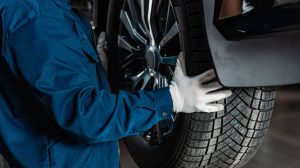 An engineer wearing blue overalls and white gloves handles a tire on a vehicle in a workshop.